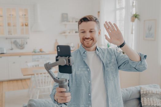 Photo of handsome guy in living room enjoying weekend, streaming live video on smartphone with gimbal stabilizer, showing new flat to his friends and waving with hand in hello gesture, casual dressed