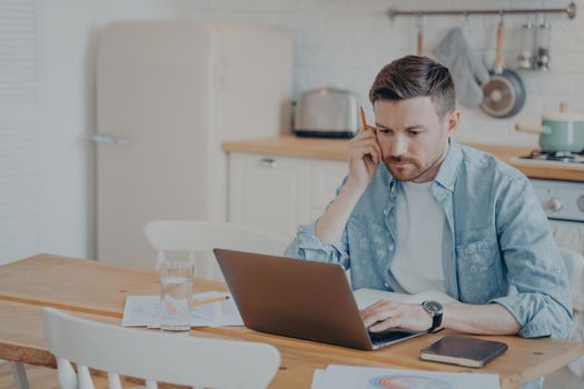 Serious focused determined young bearded businessman or freelancer in casual clothes sitting at kitchen table and typing on laptop, working on project online, researching or writing email to client