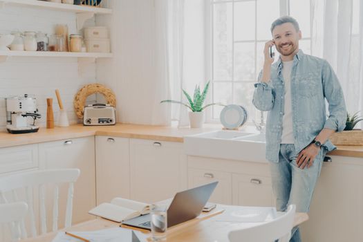 Happy young male involved in pleasant phone conversation at kitchen while working online on laptop, young man entrepreneur talking to client on cellphone and offering successful financial transaction