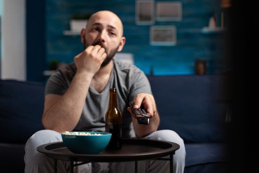 Scared shocked man looking at horror thriller movie at tv, eating popcorn trembled with fright sitting on comfortable sofa. Concentrated alone male watching tv late night enjoying free time