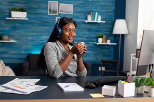 African american student holding cup of coffee in hands wearing headphones while listening modern music. Black woman working remote from home browsing school course using elearning platform