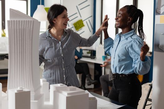 Architecture multi ethnic workmates in partnership to design building model maquette. Diverse professional engineer women highfive after success in blueprint plans cooperation