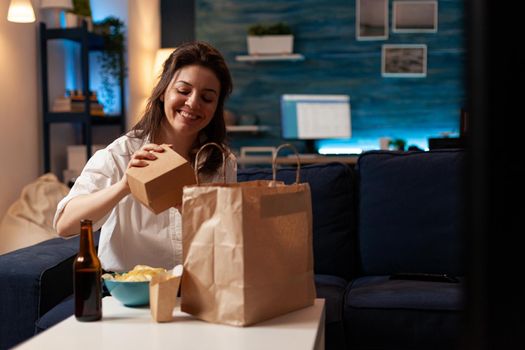 Smiling cheerful woman unpacking tasting fast-food home delivered sitting on couch having lunch serivce order. Caucasian female enjoying delicious takeaway delivery tasty meal in evening