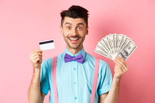 Cheerful caucasian man with moustache and bow-tie showing plastic credit card with money in dollars, smiling at camera, standing over pink background.