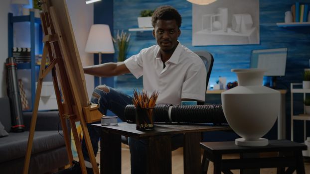 African american person drawing vase design on white canvas in creativity studio. Close up of crafting pencils on table while black young man creating masterpiece in background space