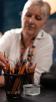 Close up of senior artist looking at colorful pencils for masterpiece drawing on table sitting in workshop space. Elder woman with artistic imagination using tools for professional art project