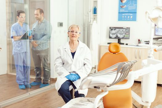 Stomatologist senior woman looking into camera while waiting for sick man in dentistry examination room. Medical nurse discussing with patient typing tooth diagnosis on clipboard