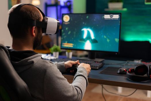 Back view of gamer playing space shooter competition using virtual reality goggles. Competitive player using joystick for online championship sitting on gamining chair late at night in living room