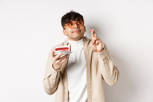 Cute hopeful guy in glasses making birthday wish, holding cake and fingers crossed, looking up and praying, hope dream come true, standing on white background.