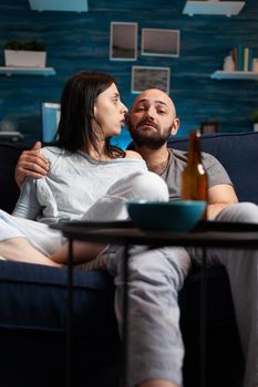 Confused astonished young couple watching documentary show having shocked facial expression, eating popcorn sitting on couch. Concentrated adults looking at tv late night enjoying free time