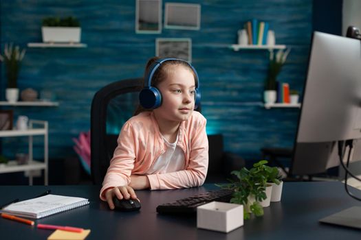 Clever school girl using computer monitor at home while wearing headphone set listening to teacher on online class meeting for education. Small student sitting with technology knowledge