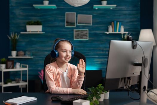 Little student waving at video call conference on computer for online math class using internet technology. Small caucasian girl with headphones and gadget studying for primary education