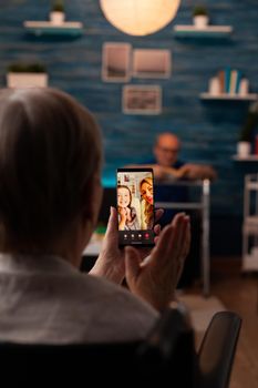 Grandmother talking to family on internet video call using online smartphone technology at home. Retired woman enjoying chat with daughter and niece while sitting in living room with grandfather