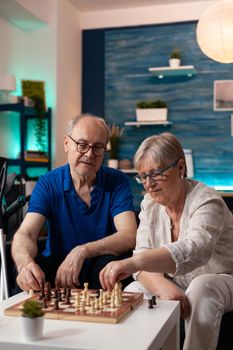 Retirement aged people sitting on living room sofa at home while playing chess game on board to relax. Caucasian old couple enjoying indoor fun activity resting on couch with crutches