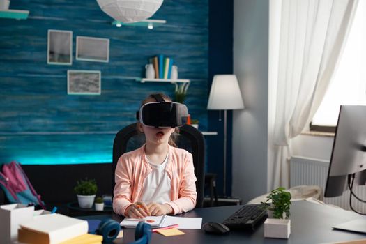 Little pupil at home desk using vr glasses for class lesson studying virtual reality knowledge. Schoolgirl with modern equipment gadget for intelligent learning educational process