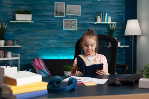 Little caucasian student holding book reading pages for school work assignment lesson on online internet class. Smart girl improving knowledge for educational progress concept and exam