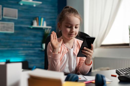 Young student waving at video call camera on smartphone while talking to teacher for online classes and elementary school homework. Small girl sitting at desk indoors with mobile phone