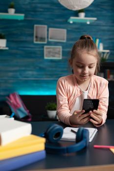 Elementary school pupil holding smartphone at desk for online communication video call conference class. Caucasian little girl using technology for studying lessons and homework