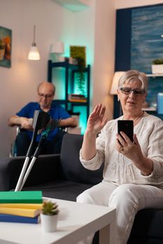 Mature retired couple on modern video call at home using smartphone technology online conference. Elder man reading while senior woman talking to family on webcam internet connection