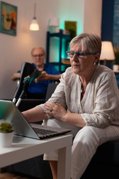 Elder caucasian people enjoying activities being at home. Old wife using modern laptop computer sitting on couch while husband in wheelchair reading. Senior mature handicapped couple