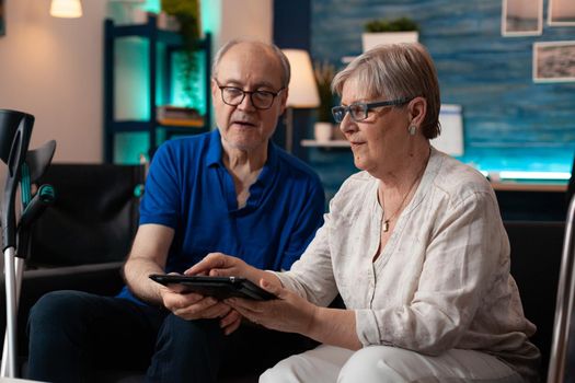 Old adult couple holding digital tablet at home on couch using modern technology for entertainment and online internet communication. Elder man and woman with crutches and walk frame