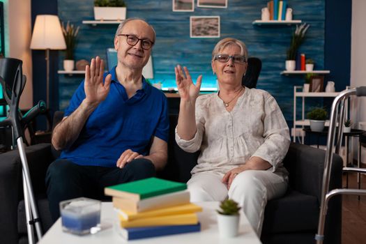 Portrait of retirement couple sitting on couch waving hands at camera in living room. Old man and woman with walking disablement having crutches and walk frame for transportation support