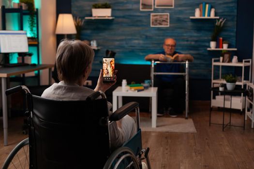 Disabled woman in wheelchair using video call technology on online internet connection smartphone talking to family. Senior people with injury sitting at home with helpful equipment