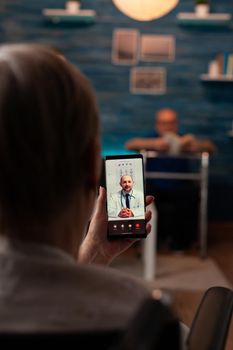 Retired woman holding smartphone with video call for doctor appointment and telemedicine consultation in living room at home. Old man with walk frame sitting on sofa reading book