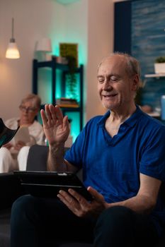 Old man waving at video call webcam on tablet device with modern technology gadget and wireless internet sitting on couch, while senior woman disabled in wheelchair reading book