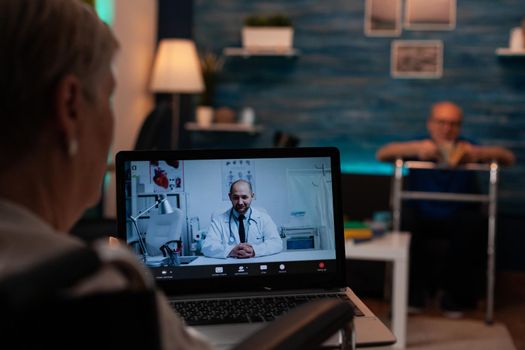 Old woman talking to doctor on video call conference for remote consultation on laptop computer to receive prescription medicine and advice. Elder man with walk frame sitting on couch