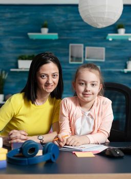 Portrait of mother and daughter sitting at home desk waiting for online class lesson in elementary school. Caucasian adult assisting young child learning for good education on internet