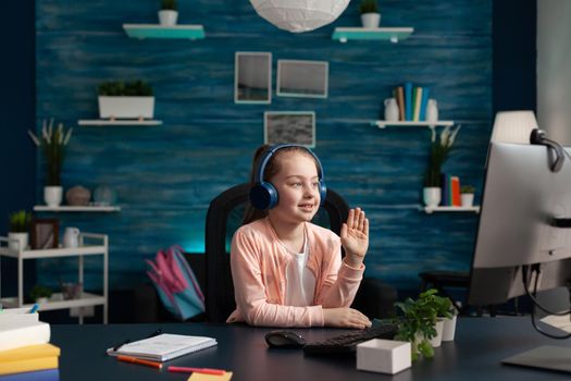 Little student waving at video call conference on computer for online math class using internet technology. Small caucasian girl with headphones and gadget studying for primary education