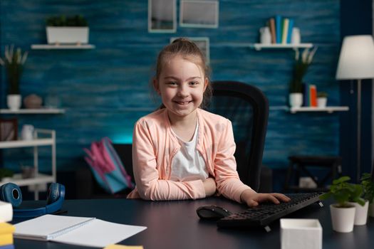 Portrait of little primary school kid sitting at home desk smiling. Caucasian pupil girl waiting for online class lesson on internet to start. Child with notebook on table for homework