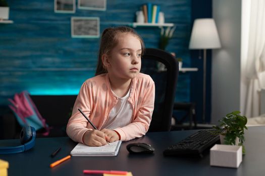 Little kid taking notes on class notebook for learning and studying lesson. Clever schoolgirl attending online meeting writing information while using internet connection for education