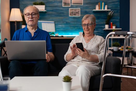 Senior family using modern technology devices laptop computer smartphone for browsing internet online while sitting on living room couch. Old people learning about digital gadget