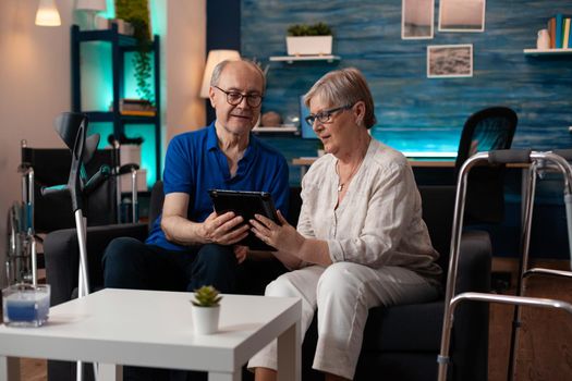 Old adult couple holding digital tablet at home on couch using modern technology for entertainment and online internet communication. Elder man and woman with crutches and walk frame