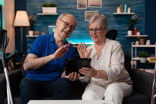 Old husband and wife waving on video call using tablet with online internet technology gadget sitting at home. Caucasian couple having virtual conference connection for communication