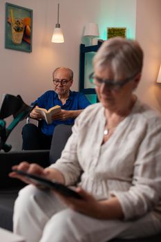 Senior retired woman looking at digital tablet gadget sitting on couch in living room while old man in background reading book. Elder couple with disability problems relaxing at home
