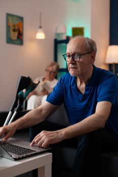 Senior elderly people enjoying retirement in living room at home. Caucasian woman in wheelchair reading and old man looking at virtual device laptop computer internet with crutches