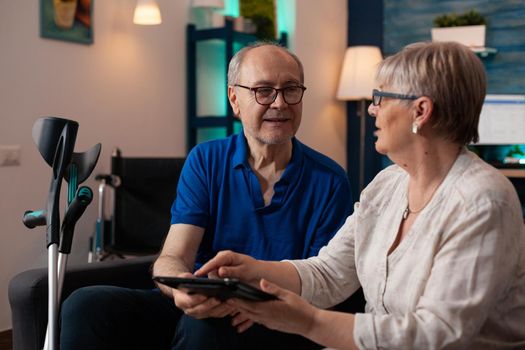 Elder couple using modern tablet with online internet on technology device in living room at home. Caucasian married man and woman with crutches and wheelchair for retirement support