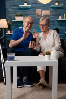 Elderly couple waving at video call webcam using smartphone technology with modern online internet connection sitting on couch. Caucasian people meeting on virtual conference with family