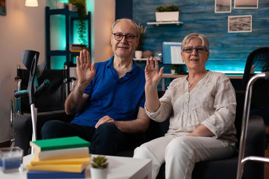Portrait of retirement couple sitting on couch waving hands at camera in living room. Old man and woman with walking disablement having crutches and walk frame for transportation support