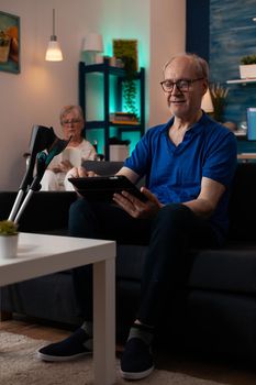 Couple of pensioners sitting at home with modern technology and healthcare support equipment. Caucasian man using digital tablet gadget on couch with crutches and woman in wheelchair