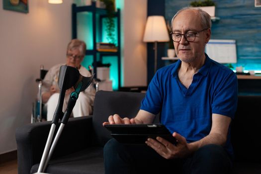 Couple of pensioners sitting at home with modern technology and healthcare support equipment. Caucasian man using digital tablet gadget on couch with crutches and woman in wheelchair