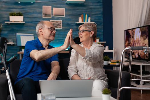 Old aged married couple high five using laptop computer on living room couch at home. Caucasian elder husband and wife with technology gadget device for digital internet entertainment