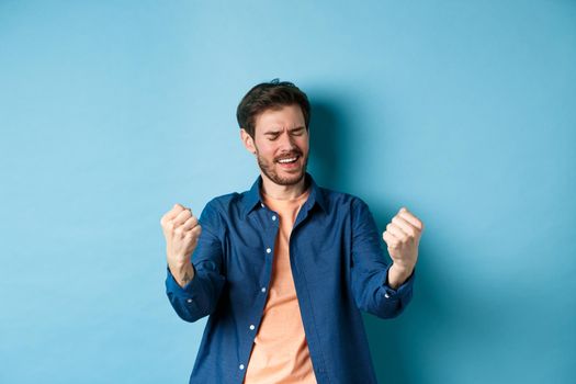 Image of happy man celebrating success, shouting yes and shaking clenched fists, achieve goal and triumphing, winning and dancing, standing on blue background.