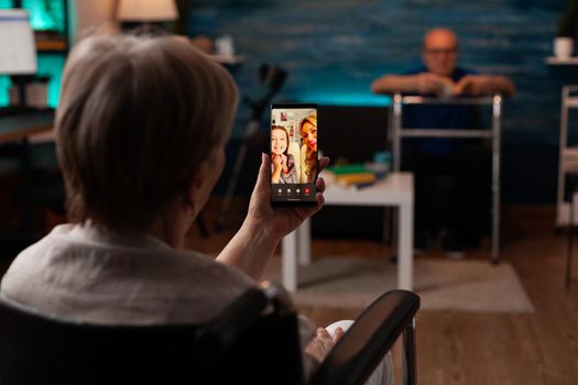 Elderly woman holding smartphone chatting to family on online video call using smartphone internet technology. Old person talking to daughter and niece sitting in living room with senior husband