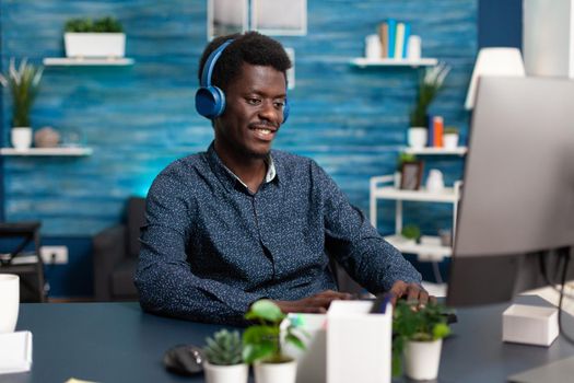 Smiling african american man using laptop with headphones working from home and listening webinar or online courses. Black student taking internet classes from home office desk