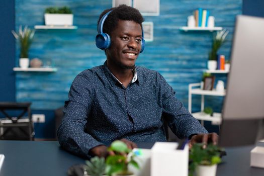 Authentic smiling african american man using laptop and headphones to work from home and learn freelancer webinar or online college courses. Business guy watching movie indoors at desk