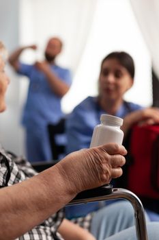 Disabled senior elderly patient holding vial of pills discussing disease therapy with support assistant worker. Woman nurse holding emergency red kit bag in hands. Social nusing services at home